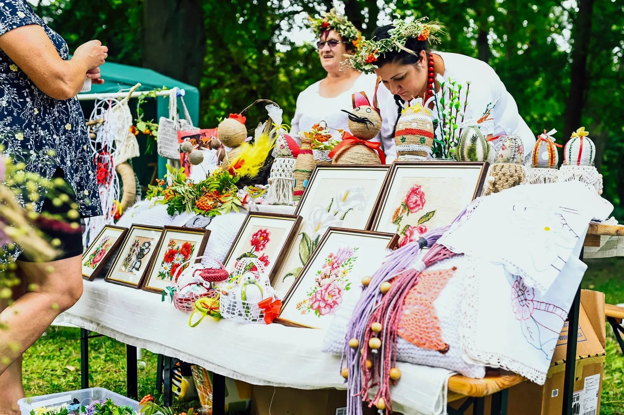 Stoisko jarmarczne z haftowanymi obrazkami, makramami i ozdobami; dwie panie z wiankami z ziół